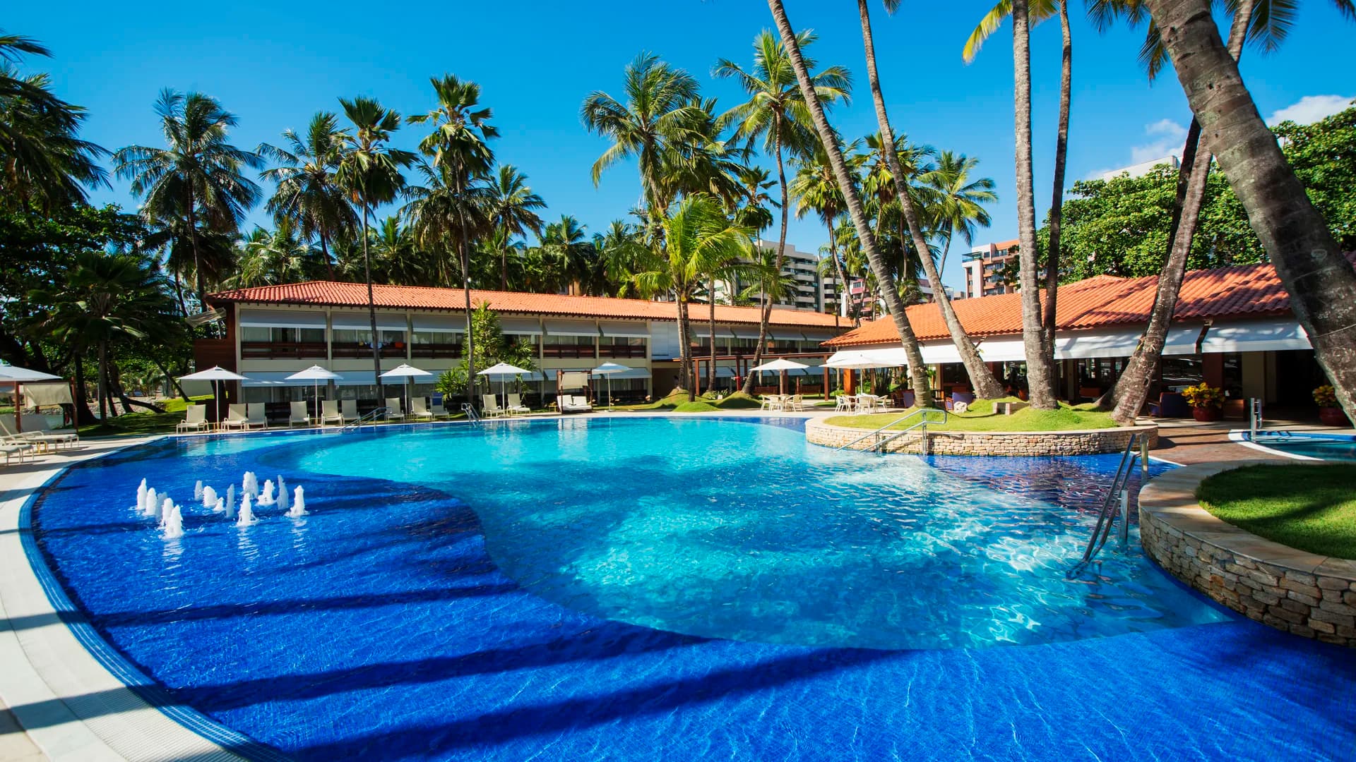 Piscina do Jatiúca com jatos de água ligados, cercada por coqueiral